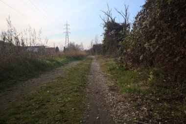 Elektrik direği ve sınırında bir elektrik hattı olan patika, gün batımında İtalya 'nın kırsal kesimindeki bir parkta, bir köyün evleriyle sınırda, ve eğlence kanalının diğer tarafında bir viyadük kanalı ile çevrili.