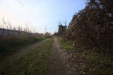 Elektrik direği ve sınırında bir elektrik hattı olan patika, gün batımında İtalya 'nın kırsal kesimindeki bir parkta, bir köyün evleriyle sınırda, ve eğlence kanalının diğer tarafında bir viyadük kanalı ile çevrili.