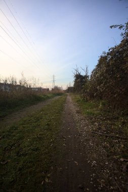 Elektrik direği ve sınırında bir elektrik hattı olan patika, gün batımında İtalya 'nın kırsal kesimindeki bir parkta, bir köyün evleriyle sınırda, ve eğlence kanalının diğer tarafında bir viyadük kanalı ile çevrili.