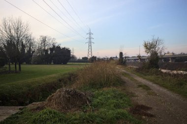 Elektrik direği ve sınırında bir elektrik hattı olan patika, gün batımında İtalya 'nın kırsal kesimindeki bir parkta, bir köyün evleriyle sınırda, ve eğlence kanalının diğer tarafında bir viyadük kanalı ile çevrili.