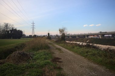 Elektrik direği ve sınırında bir elektrik hattı olan patika, gün batımında İtalya 'nın kırsal kesimindeki bir parkta, bir köyün evleriyle sınırda, ve eğlence kanalının diğer tarafında bir viyadük kanalı ile çevrili.