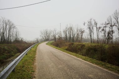 Kışın İtalya kırsalında sisli bir günde, bir su deresi, çıplak ağaçlar ve uzun otlarla çevrili bir viraj yaparak setin üzerindeki yola tırmanmak.