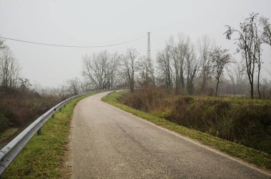 Kışın İtalya kırsalında sisli bir günde, bir su deresi, çıplak ağaçlar ve uzun otlarla çevrili bir viraj yaparak setin üzerindeki yola tırmanmak.
