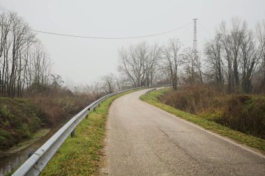 Kışın İtalya kırsalında sisli bir günde, bir su deresi, çıplak ağaçlar ve uzun otlarla çevrili bir viraj yaparak setin üzerindeki yola tırmanmak.