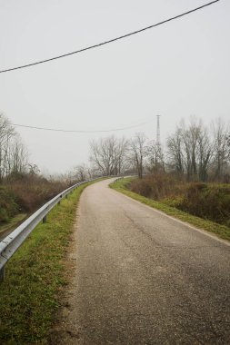 Kışın İtalya kırsalında sisli bir günde, bir su deresi, çıplak ağaçlar ve uzun otlarla çevrili bir viraj yaparak setin üzerindeki yola tırmanmak.