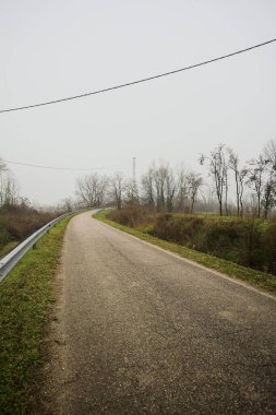 Kışın İtalya kırsalında sisli bir günde, bir su deresi, çıplak ağaçlar ve uzun otlarla çevrili bir viraj yaparak setin üzerindeki yola tırmanmak.