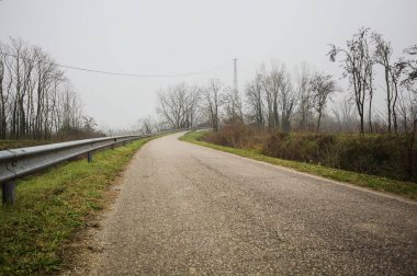 Kışın İtalya kırsalında sisli bir günde, bir su deresi, çıplak ağaçlar ve uzun otlarla çevrili bir viraj yaparak setin üzerindeki yola tırmanmak.