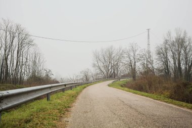 Kışın İtalya kırsalında sisli bir günde, bir su deresi, çıplak ağaçlar ve uzun otlarla çevrili bir viraj yaparak setin üzerindeki yola tırmanmak.