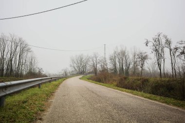 Kışın İtalya kırsalında sisli bir günde, bir su deresi, çıplak ağaçlar ve uzun otlarla çevrili bir viraj yaparak setin üzerindeki yola tırmanmak.