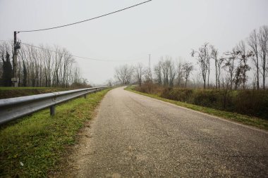 Kışın İtalya kırsalında sisli bir günde, bir su deresi, çıplak ağaçlar ve uzun otlarla çevrili bir viraj yaparak setin üzerindeki yola tırmanmak.