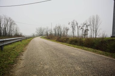 Kışın İtalya kırsalında sisli bir günde, bir su deresi, çıplak ağaçlar ve uzun otlarla çevrili bir viraj yaparak setin üzerindeki yola tırmanmak.