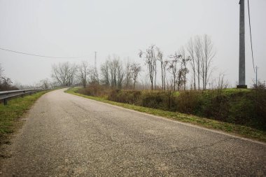 Kışın İtalya kırsalında sisli bir günde, bir su deresi, çıplak ağaçlar ve uzun otlarla çevrili bir viraj yaparak setin üzerindeki yola tırmanmak.