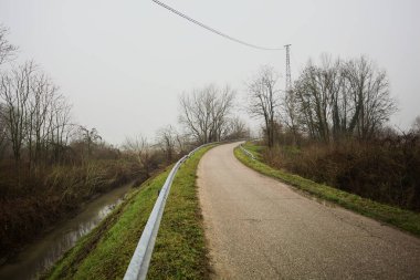 Kışın İtalya kırsalında sisli bir günde, bir su deresi, çıplak ağaçlar ve uzun otlarla çevrili bir viraj yaparak setin üzerindeki yola tırmanmak.
