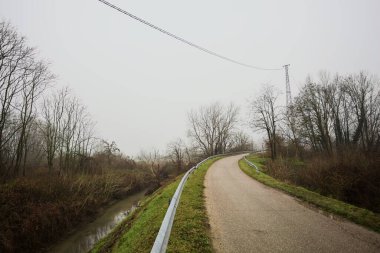 Kışın İtalya kırsalında sisli bir günde, bir su deresi, çıplak ağaçlar ve uzun otlarla çevrili bir viraj yaparak setin üzerindeki yola tırmanmak.