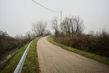 Kışın İtalya kırsalında sisli bir günde, bir su deresi, çıplak ağaçlar ve uzun otlarla çevrili bir viraj yaparak setin üzerindeki yola tırmanmak.