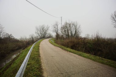 Kışın İtalya kırsalında sisli bir günde, bir su deresi, çıplak ağaçlar ve uzun otlarla çevrili bir viraj yaparak setin üzerindeki yola tırmanmak.