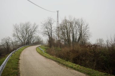 Kışın İtalya kırsalında sisli bir günde, bir su deresi, çıplak ağaçlar ve uzun otlarla çevrili bir viraj yaparak setin üzerindeki yola tırmanmak.