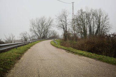 Kışın İtalya kırsalında sisli bir günde, bir su deresi, çıplak ağaçlar ve uzun otlarla çevrili bir viraj yaparak setin üzerindeki yola tırmanmak.