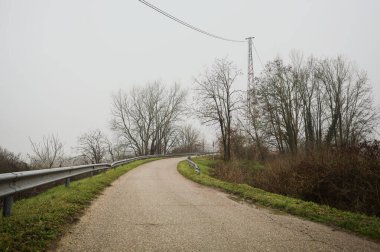 Kışın İtalya kırsalında sisli bir günde, bir su deresi, çıplak ağaçlar ve uzun otlarla çevrili bir viraj yaparak setin üzerindeki yola tırmanmak.