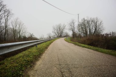 Kışın İtalya kırsalında sisli bir günde, bir su deresi, çıplak ağaçlar ve uzun otlarla çevrili bir viraj yaparak setin üzerindeki yola tırmanmak.