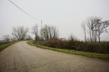 Kışın İtalya kırsalında sisli bir günde, bir su deresi, çıplak ağaçlar ve uzun otlarla çevrili bir viraj yaparak setin üzerindeki yola tırmanmak.