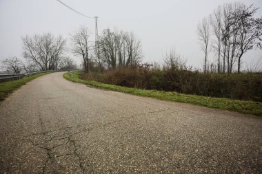 Kışın İtalya kırsalında sisli bir günde, bir su deresi, çıplak ağaçlar ve uzun otlarla çevrili bir viraj yaparak setin üzerindeki yola tırmanmak.
