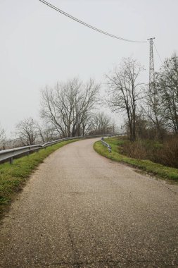Kışın İtalya kırsalında sisli bir günde, bir su deresi, çıplak ağaçlar ve uzun otlarla çevrili bir viraj yaparak setin üzerindeki yola tırmanmak.