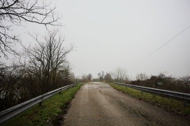 Sisli ve bulutlu bir günde, İtalya kırsalında köprünün diğer ucunda, arka planda bir malikane bulunan çıplak ağaçlar tarafından çerçevelenmiş kısmen yıpranmış yol köprüsü.
