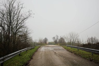 Sisli ve bulutlu bir günde, İtalya kırsalında köprünün diğer ucunda, arka planda bir malikane bulunan çıplak ağaçlar tarafından çerçevelenmiş kısmen yıpranmış yol köprüsü.