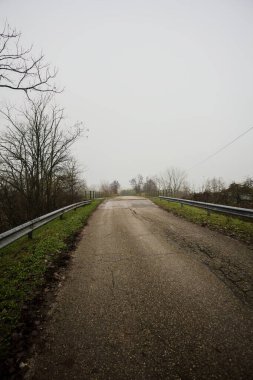 Sisli ve bulutlu bir günde, İtalya kırsalında köprünün diğer ucunda, arka planda bir malikane bulunan çıplak ağaçlar tarafından çerçevelenmiş kısmen yıpranmış yol köprüsü.