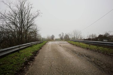 Sisli ve bulutlu bir günde, İtalya kırsalında köprünün diğer ucunda, arka planda bir malikane bulunan çıplak ağaçlar tarafından çerçevelenmiş kısmen yıpranmış yol köprüsü.