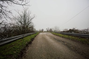 Sisli ve bulutlu bir günde, İtalya kırsalında köprünün diğer ucunda, arka planda bir malikane bulunan çıplak ağaçlar tarafından çerçevelenmiş kısmen yıpranmış yol köprüsü.