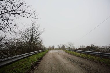 Sisli ve bulutlu bir günde, İtalya kırsalında köprünün diğer ucunda, arka planda bir malikane bulunan çıplak ağaçlar tarafından çerçevelenmiş kısmen yıpranmış yol köprüsü.