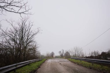 Sisli ve bulutlu bir günde, İtalya kırsalında köprünün diğer ucunda, arka planda bir malikane bulunan çıplak ağaçlar tarafından çerçevelenmiş kısmen yıpranmış yol köprüsü.