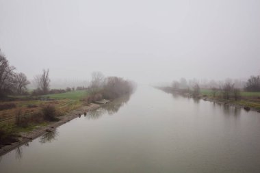 İtalya 'nın kırsal kesimlerinde sisli ve bulutlu bir günde sulara yansıyan ağaçlarla çevrili tarlalar arasındaki nehir.