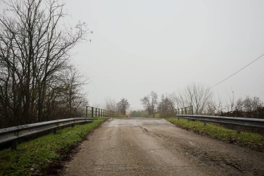 Sisli ve bulutlu bir günde, İtalya kırsalında köprünün diğer ucunda, arka planda bir malikane bulunan çıplak ağaçlar tarafından çerçevelenmiş kısmen yıpranmış yol köprüsü.