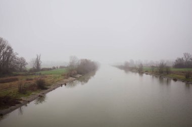 İtalya 'nın kırsal kesimlerinde sisli ve bulutlu bir günde sulara yansıyan ağaçlarla çevrili tarlalar arasındaki nehir.
