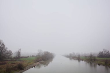 İtalya 'nın kırsal kesimlerinde sisli ve bulutlu bir günde sulara yansıyan ağaçlarla çevrili tarlalar arasındaki nehir.