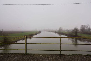 Sisli bir günde, İtalya 'nın kırsal kesiminde ağaçlar suya dökülen nehir, bir köprüden görülüyor ve korkuluklarıyla çerçeveleniyor.