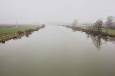 İtalya 'nın kırsal kesimlerinde sisli ve bulutlu bir günde sulara yansıyan ağaçlarla çevrili tarlalar arasındaki nehir.