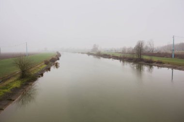 İtalya 'nın kırsal kesimlerinde sisli ve bulutlu bir günde sulara yansıyan ağaçlarla çevrili tarlalar arasındaki nehir.