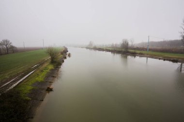 İtalya 'nın kırsal kesimlerinde sisli ve bulutlu bir günde sulara yansıyan ağaçlarla çevrili tarlalar arasındaki nehir.