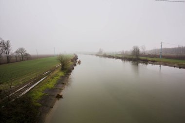 İtalya 'nın kırsal kesimlerinde sisli ve bulutlu bir günde sulara yansıyan ağaçlarla çevrili tarlalar arasındaki nehir.
