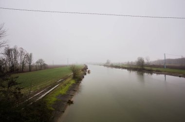 İtalya 'nın kırsal kesimlerinde sisli ve bulutlu bir günde sulara yansıyan ağaçlarla çevrili tarlalar arasındaki nehir.