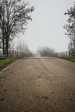 Sisli ve bulutlu bir günde, İtalya kırsalında köprünün diğer ucunda, arka planda bir malikane bulunan çıplak ağaçlar tarafından çerçevelenmiş kısmen yıpranmış yol köprüsü.