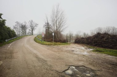 Çamurlu bir yolda yol ayrımı, bir köprüye götürür, ve ağaçlarla çevrili bir nehre uzanan çamurlu bir patika, İtalya kırsalında sisli ve bulutlu bir günde