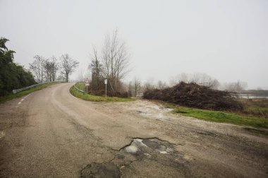 Çamurlu bir yolda yol ayrımı, bir köprüye götürür, ve ağaçlarla çevrili bir nehre uzanan çamurlu bir patika, İtalya kırsalında sisli ve bulutlu bir günde