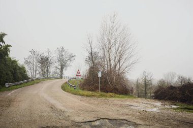Çamurlu bir yolda yol ayrımı, bir köprüye götürür, ve ağaçlarla çevrili bir nehre uzanan çamurlu bir patika, İtalya kırsalında sisli ve bulutlu bir günde