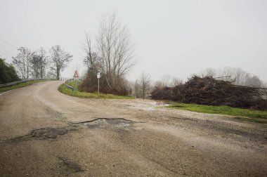 Çamurlu bir yolda yol ayrımı, bir köprüye götürür, ve ağaçlarla çevrili bir nehre uzanan çamurlu bir patika, İtalya kırsalında sisli ve bulutlu bir günde