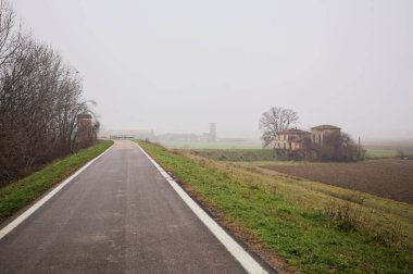 Bir toprak setin üzerindeki yol, bir tarla ile bir koruluk arasında, yolun aşağısında bir tarla, yolun kenarında tuğla bir kulübe, yolun aşağısında çıplak ağaçlarla dolu bir havza, kışın İtalya kırsalında sisli bir günde